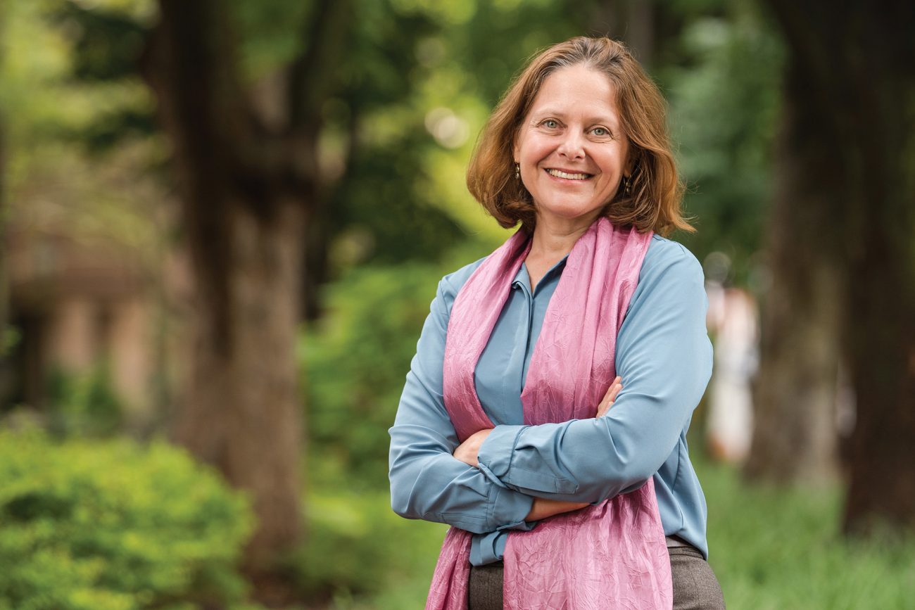 Portrait of a woman with medium-length brown hair outdoors, smiling confidently at the camera. She is dressed in a light blue shirt and a pink scarf, with arms crossed. The background is a softly blurred green environment, suggesting a serene park or garden setting.