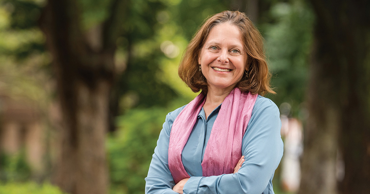 Portrait of a woman with medium-length brown hair outdoors, smiling confidently at the camera. She is dressed in a light blue shirt and a pink scarf, with arms crossed. The background is a softly blurred green environment, suggesting a serene park or garden setting.