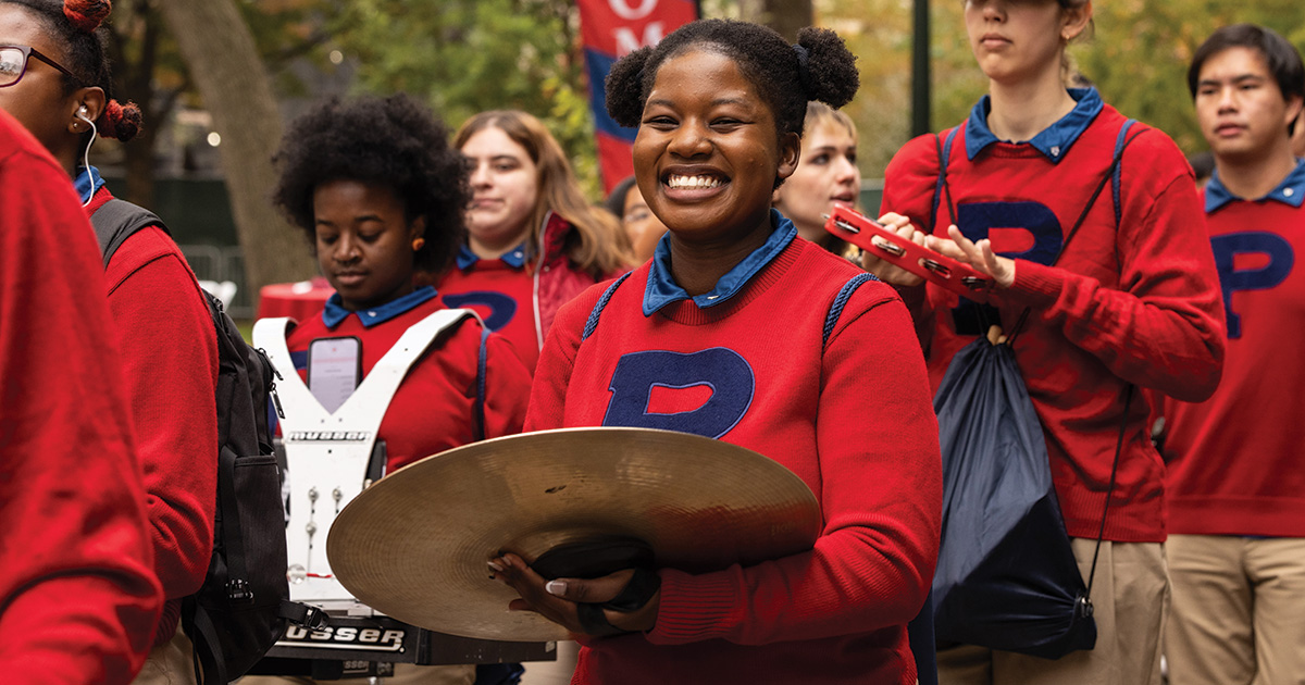 Outdoor scene of a Penn Band parade with diverse students. A young individual in the foreground, wearing a red sweater with a blue 'P,' smiles joyfully while holding a cymbal. Other band members in similar attire carry instruments like snare drums and piccolos, contributing to the lively atmosphere.