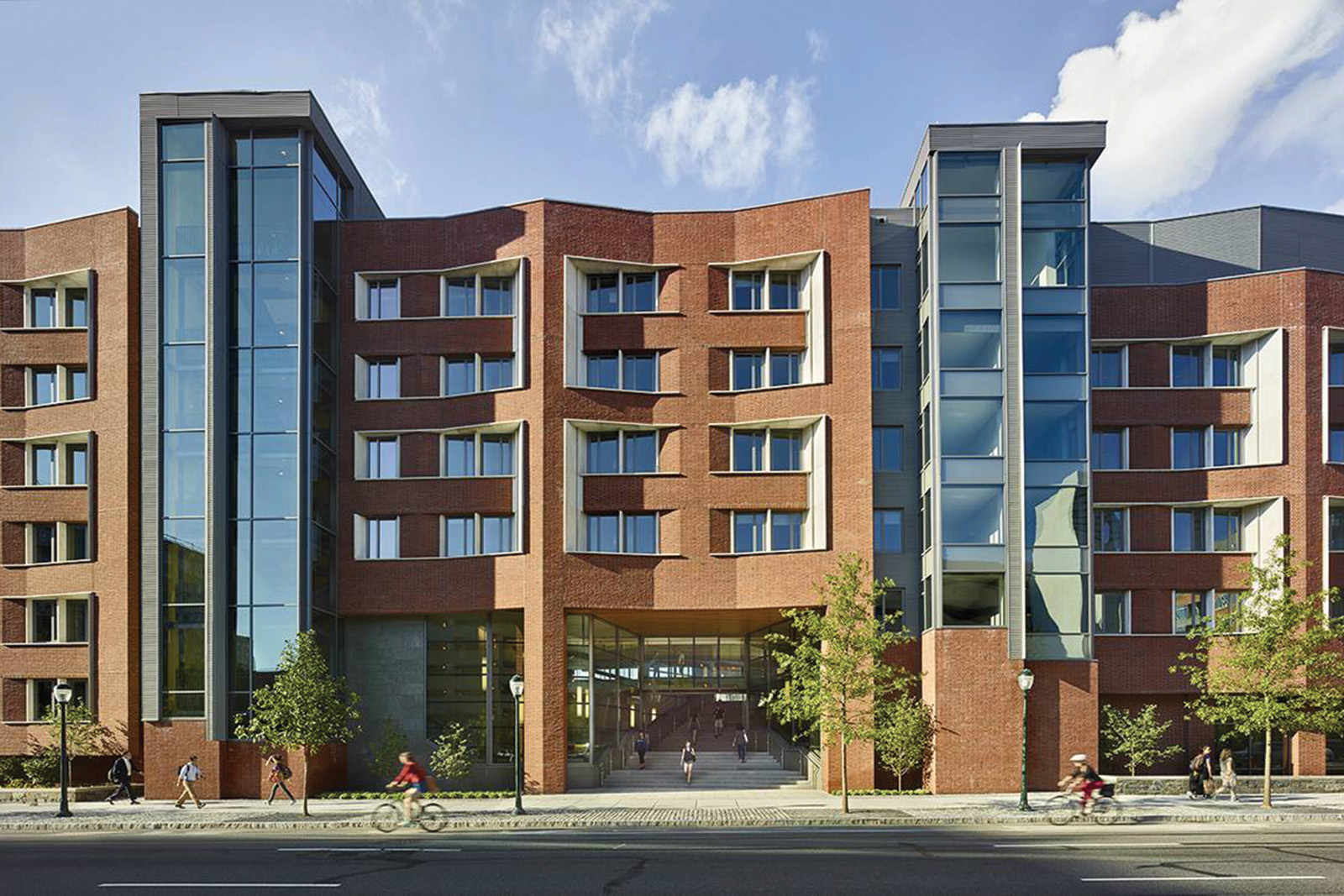 Modern urban building with a combination of brick and glass architecture. A prominent glass elevator shaft in the center adds a contemporary touch to the traditional brick sections. The scene is lively with pedestrians and a cyclist moving along the street in front, under a clear blue sky.