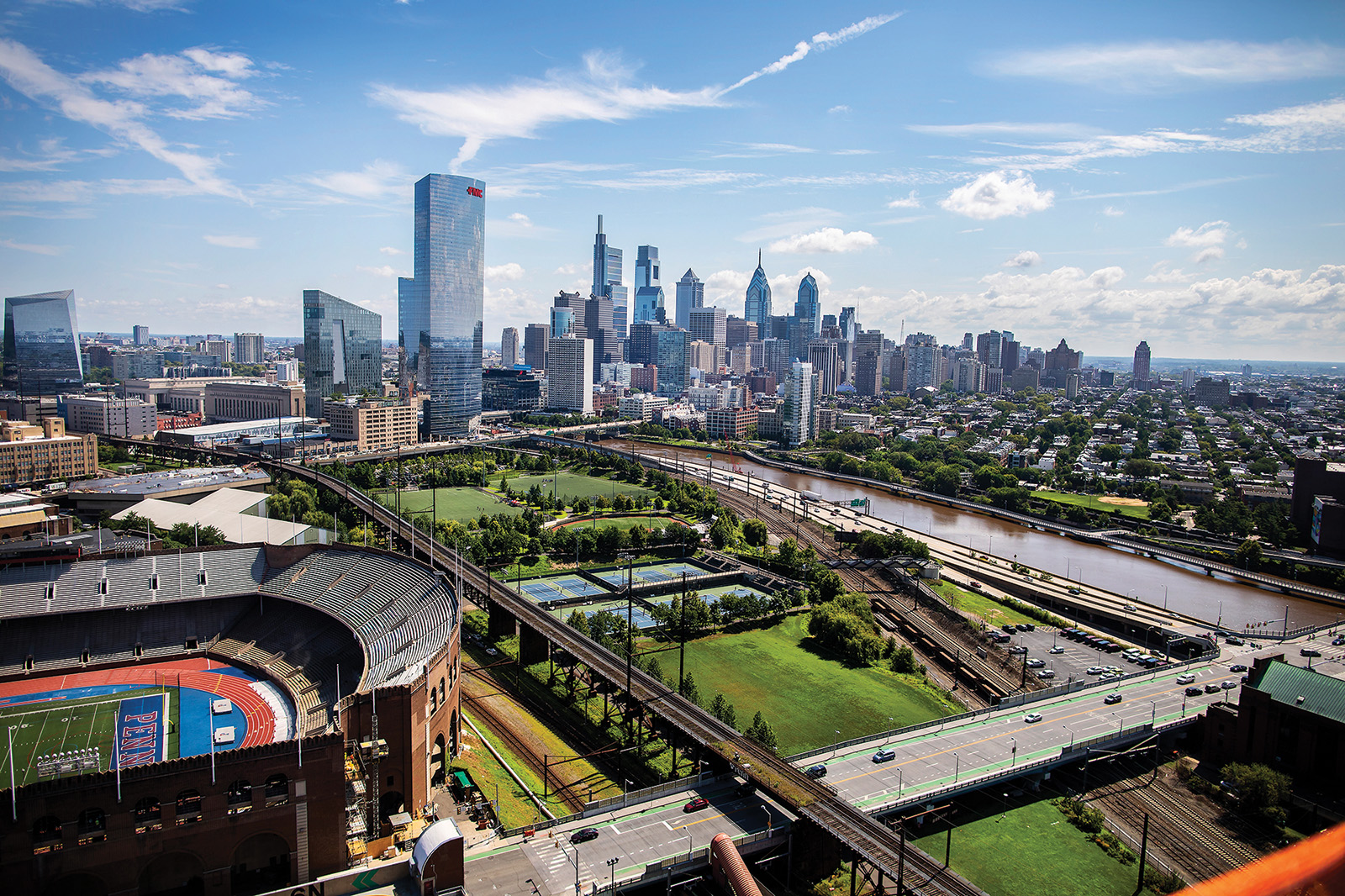 Panoramic cityscape view under a clear blue sky, featuring a sports stadium and a lush green park by a river in the foreground. The scene includes roads, railway tracks, modern glass buildings, and a skyline of tall skyscrapers, showcasing a blend of urban development and green spaces.