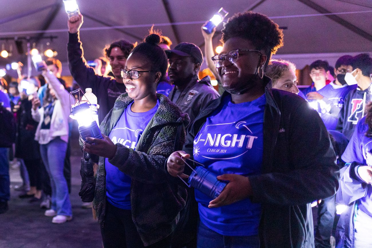 Joyful nighttime outdoor gathering with individuals participating in a class event. Two people in the foreground smile brightly and hold lanterns, lighting up the scene. They wear matching blue t-shirts with 'U-Night Class of 24'. The environment is festive with a sense of community engagement.