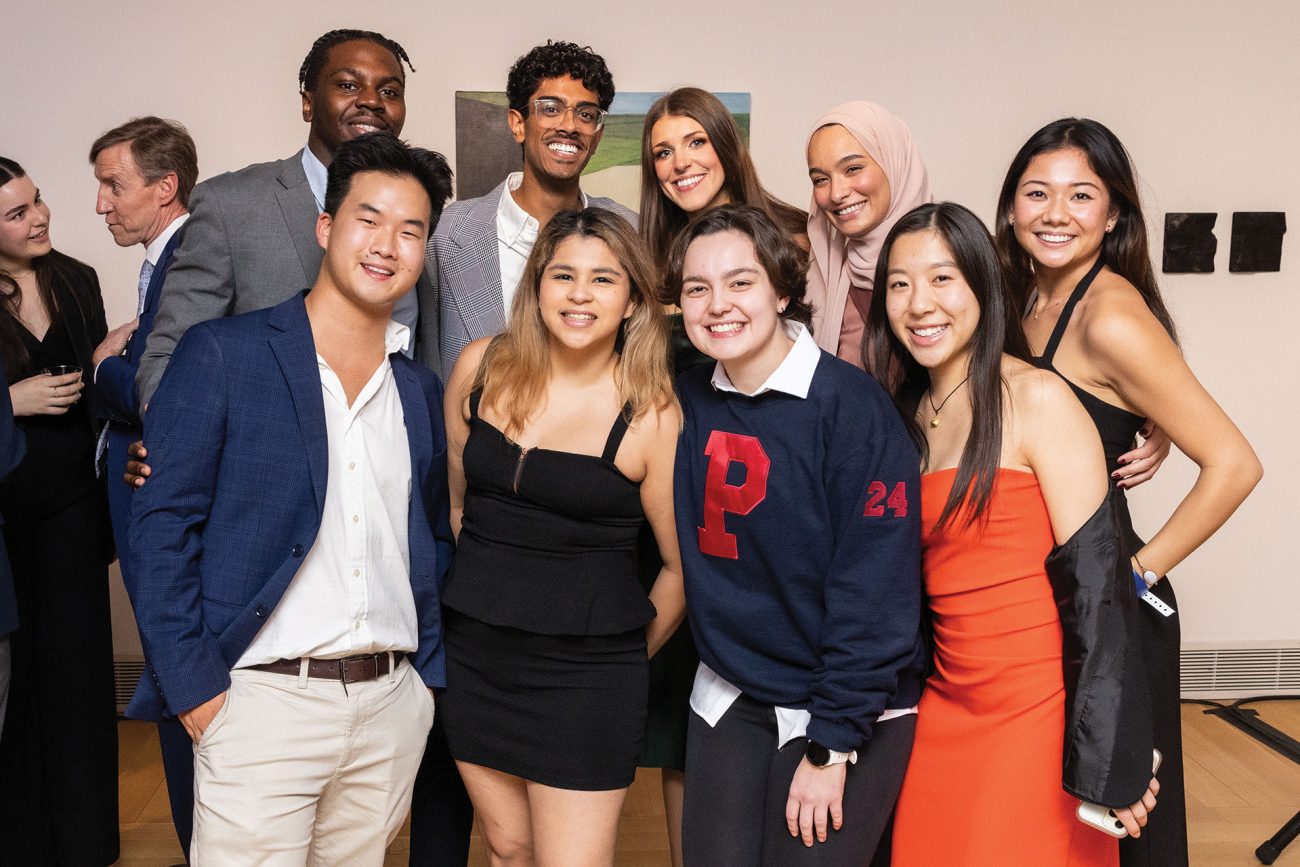 Group photo of nine diverse individuals at a semi-formal gathering, indoors. They wear a mix of styles, from suits to casual sweaters, all smiling and enjoying the event. The backdrop features subtle artwork, adding to the ambiance of the venue.