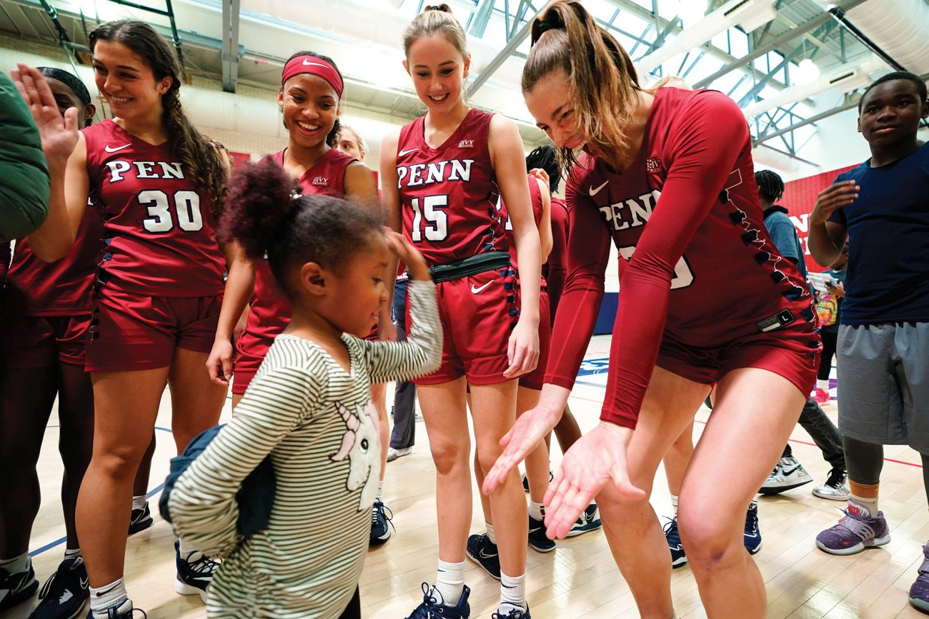 Heartwarming scene on an indoor basketball court where members of the women's basketball team in red 'PENN' uniforms engage with a young child. The players are smiling and focused on the child, who seems excited and active. The gymnasium is bustling with other participants, conveying a sense of community and inclusivity.