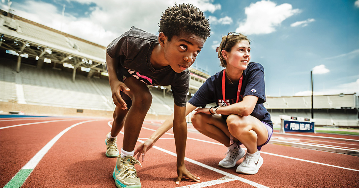 Outdoor track and field coaching session in a large stadium. A young athlete is in a starting position on the track, focusing intently ahead. A coach, beside the athlete, points to the track, offering guidance. The setting conveys a serious athletic event under a clear sky, emphasizing a supportive coaching moment.
