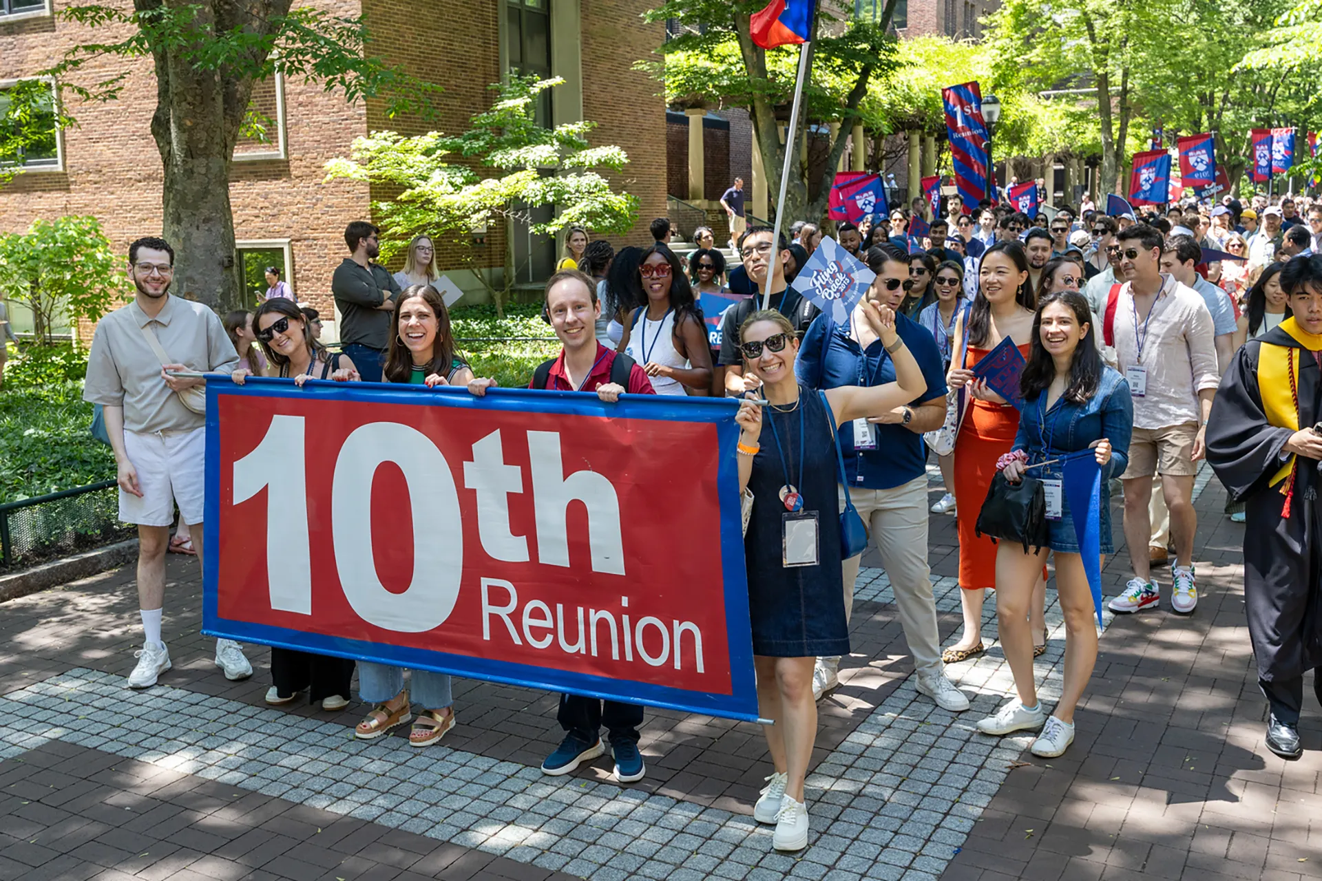 A group of young alumni holds a large blue-and-red “10th Reunion” banner as they march down a shaded brick pathway during Alumni Weekend.