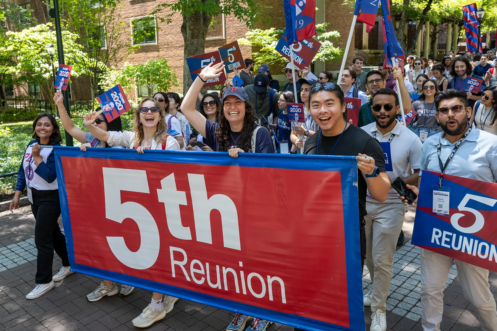 A group of young alumni marches holding a large blue-and-red “5th Reunion” banner along Locust Walk during Alumni Weekend.