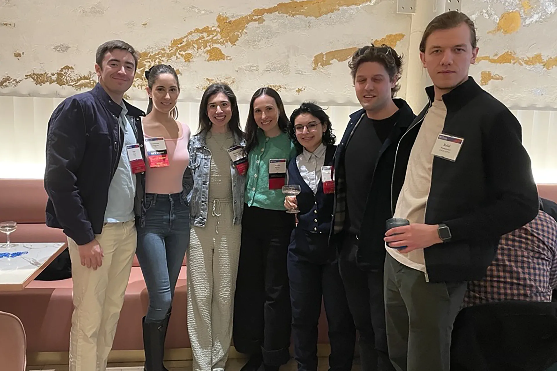 Seven smiling young alumni in name badges stand side by side at an indoor reception, some holding glasses and cups.