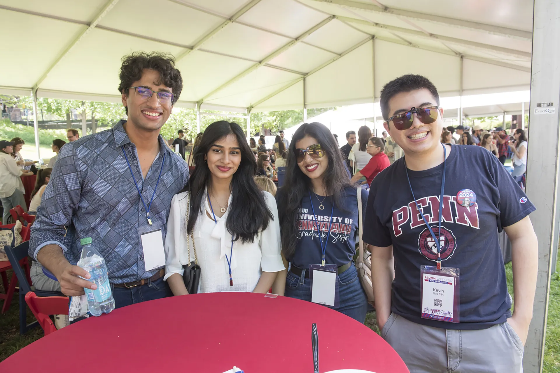 Four friends at Penn Alumni Weekend stand in front of a tented area, wearing event badges and sipping bottled water.