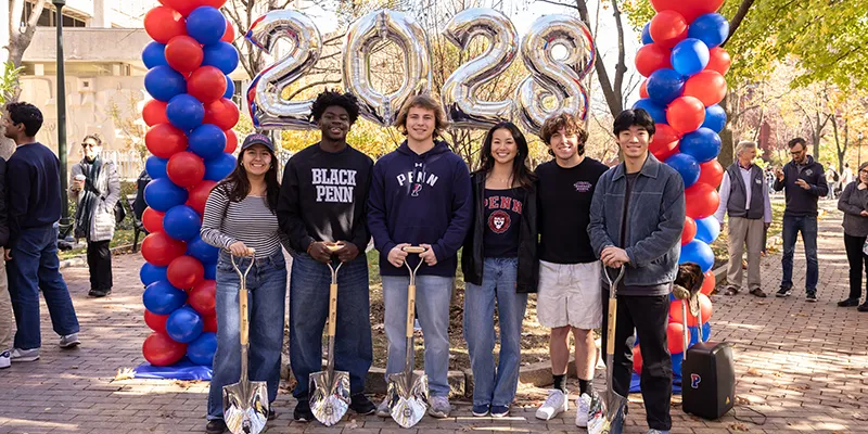 A group of Penn students from the Class of 2028 pose with shovels at the Class Tree Planting ceremony during Homecoming, framed by red and blue balloons.