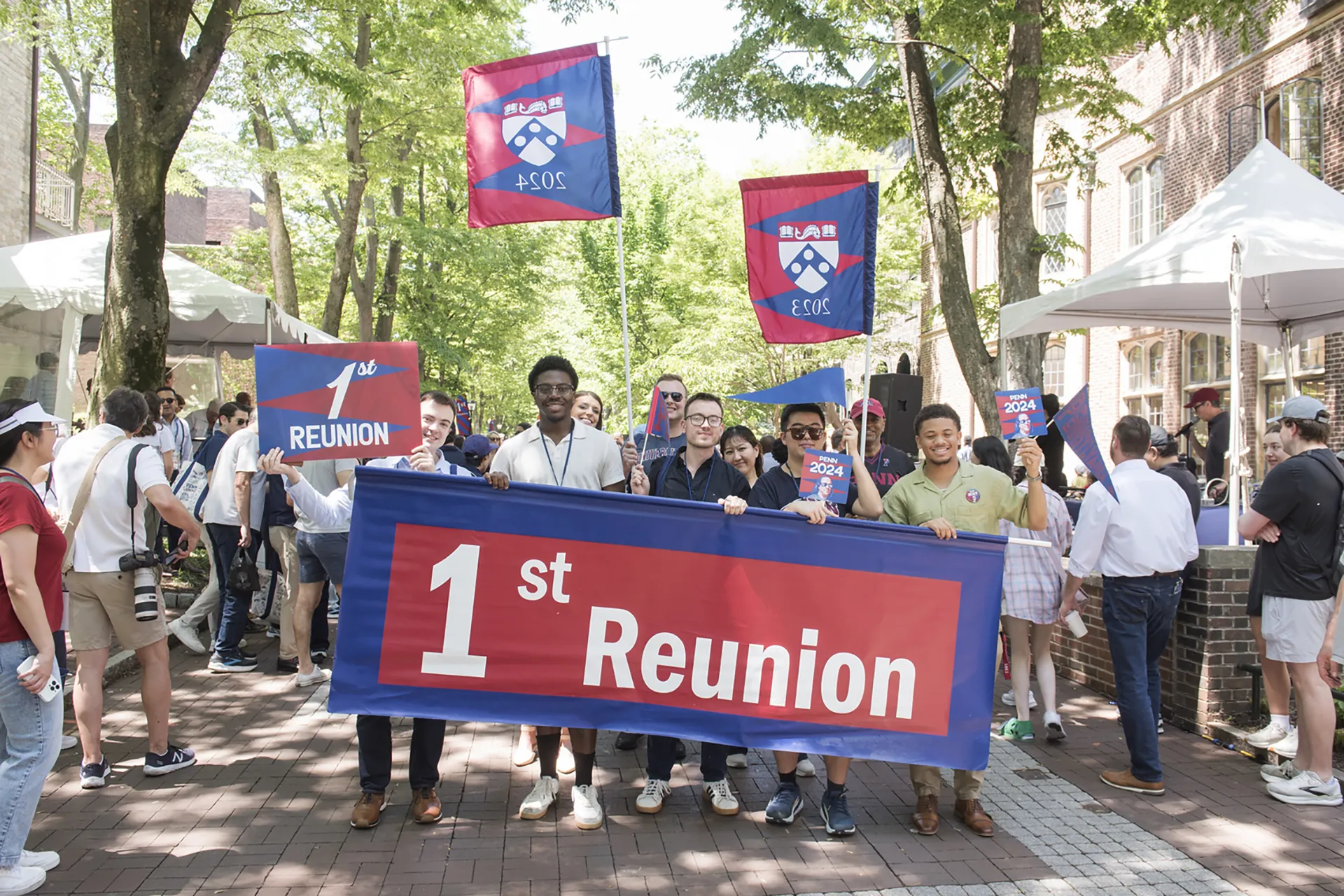 Young alumni stand beneath campus trees holding a blue-and-red “1st Reunion” banner and Penn 2024 flags during Alumni Weekend.
