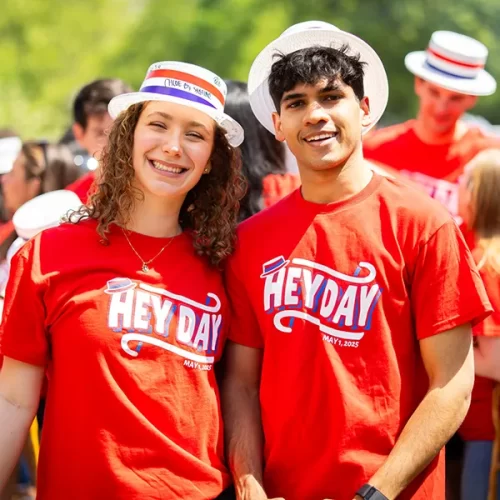 Two smiling Penn students wearing red Hey Day shirts and white hats pose for a photo during the 2025 celebration.