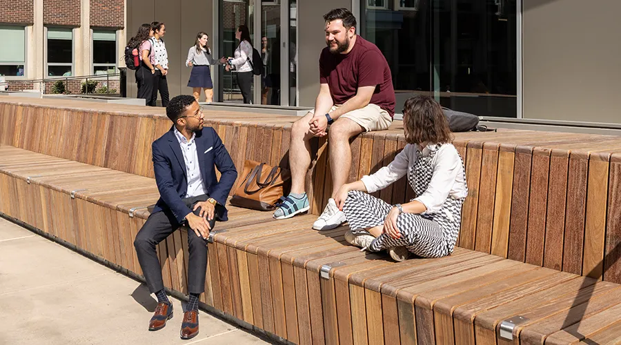 Students sit and talk outside Penn’s Graduate School of Education on a sunny day, engaging in conversation on wooden seating steps.
