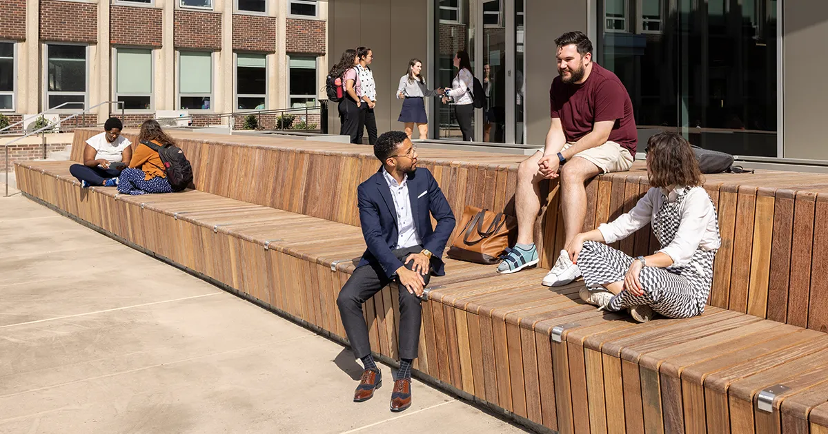 Students sit and talk outside Penn’s Graduate School of Education on a sunny day, engaging in conversation on wooden seating steps.