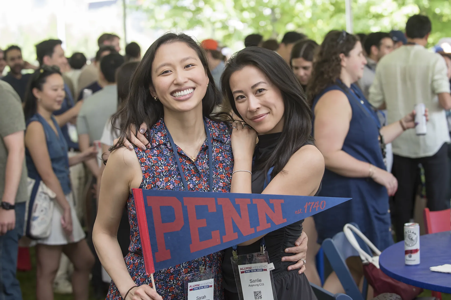 Two young alumni hug and smile under a canopy, holding a blue-and-red Penn pennant at Alumni Weekend.