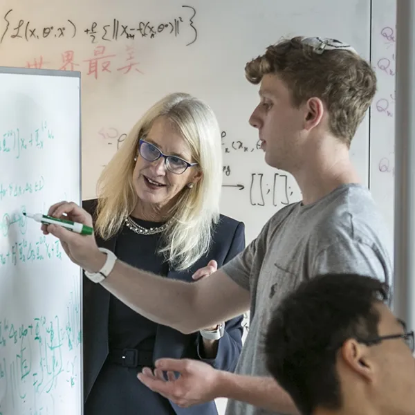 A professor in a black blazer and glasses engages with a student writing mathematical equations on a whiteboard.
