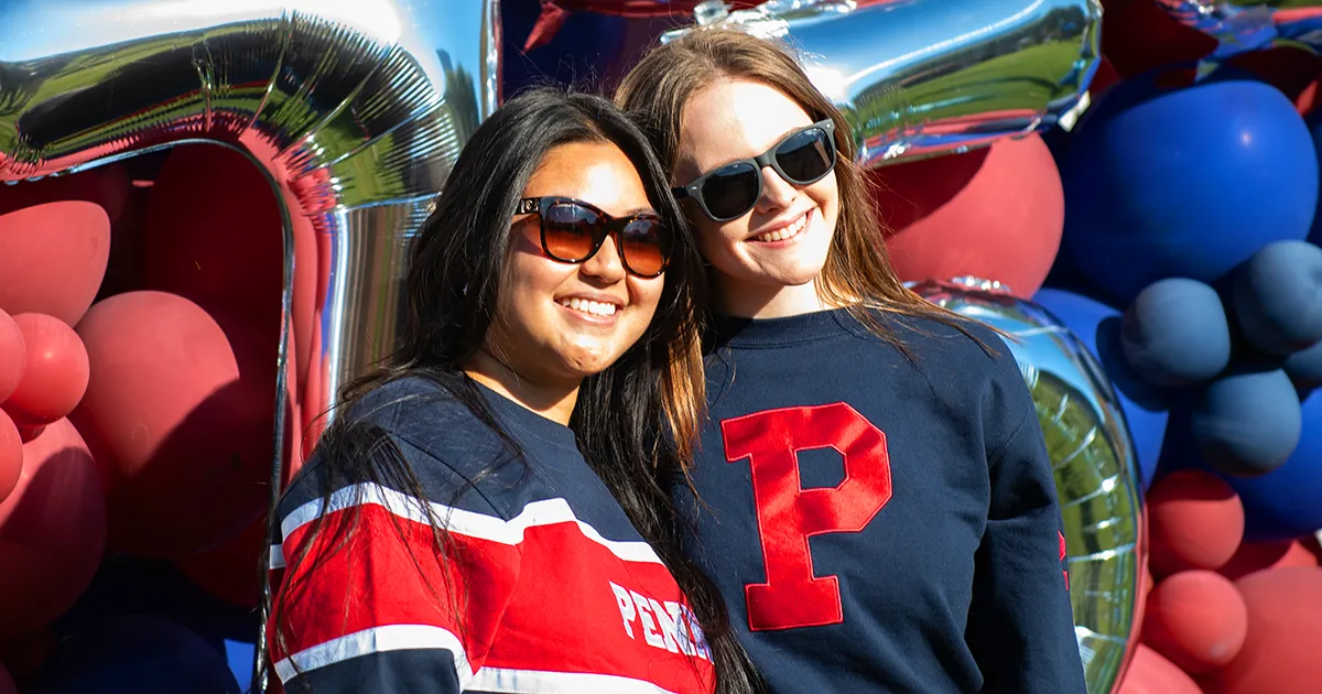 Two smiling Penn students pose in front of a red and blue balloon backdrop with metallic balloons at an event on campus.