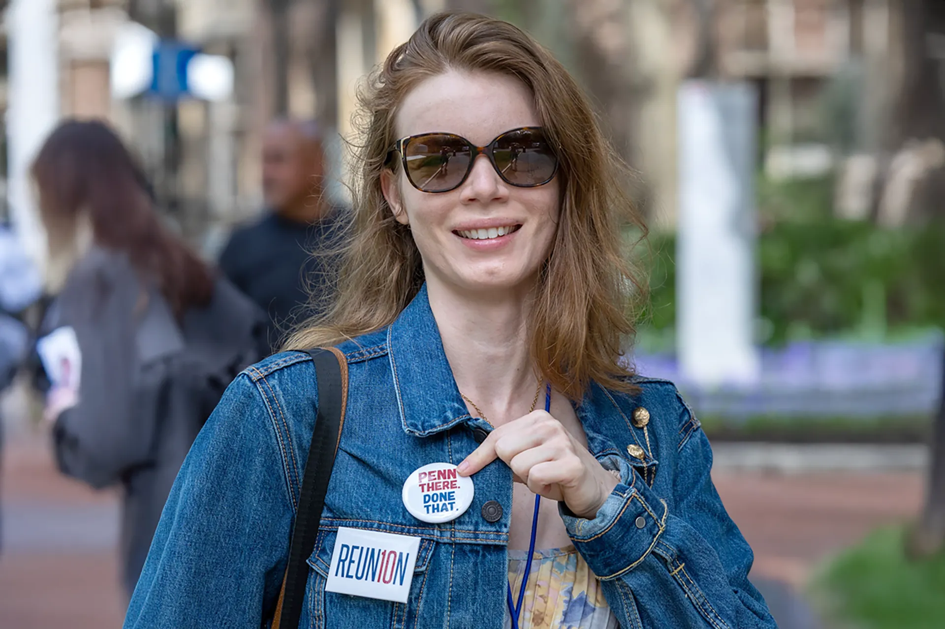 Smiling alumna in a denim jacket and sunglasses pointing to a “PENN there. DONE that.” pin with a “REUNION” sticker at Alumni Weekend.