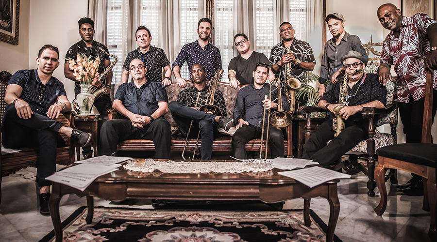 Group of twelve musicians posing in a traditional, elegant room with brass instruments, including saxophones and trombones. They are arranged in two rows with some seated on vintage furniture and others standing. The setting features floral curtains and ornate decor, suggesting a formal yet casual musical gathering.