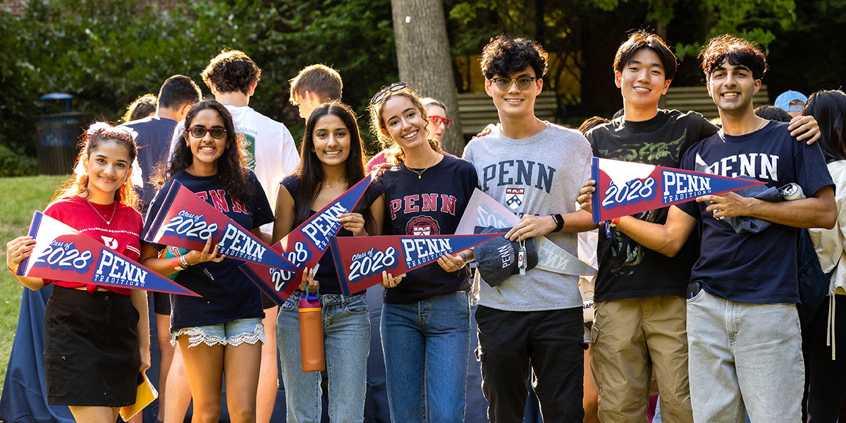 A group of University of Pennsylvania students posing outdoors at a campus event, smiling and holding pennants and graduation caps that read 