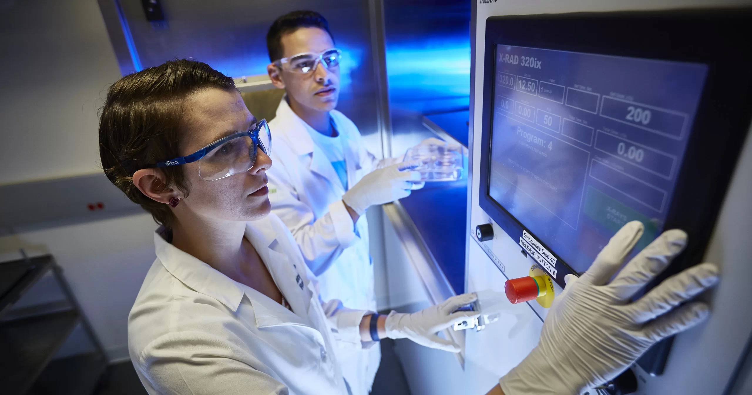 Two scientists in lab coats, safety goggles, and gloves operate a touchscreen control panel on a high-tech machine labeled 