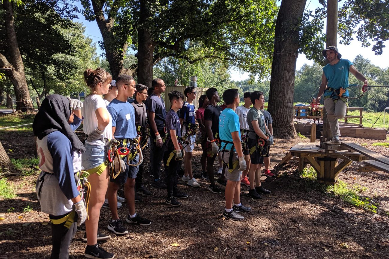 A diverse group of students, equipped with safety harnesses, attentively watch an instructor on a wooden platform demonstrating rope techniques in an outdoor park. The lush greenery and towering trees provide a natural backdrop for this team-building exercise.