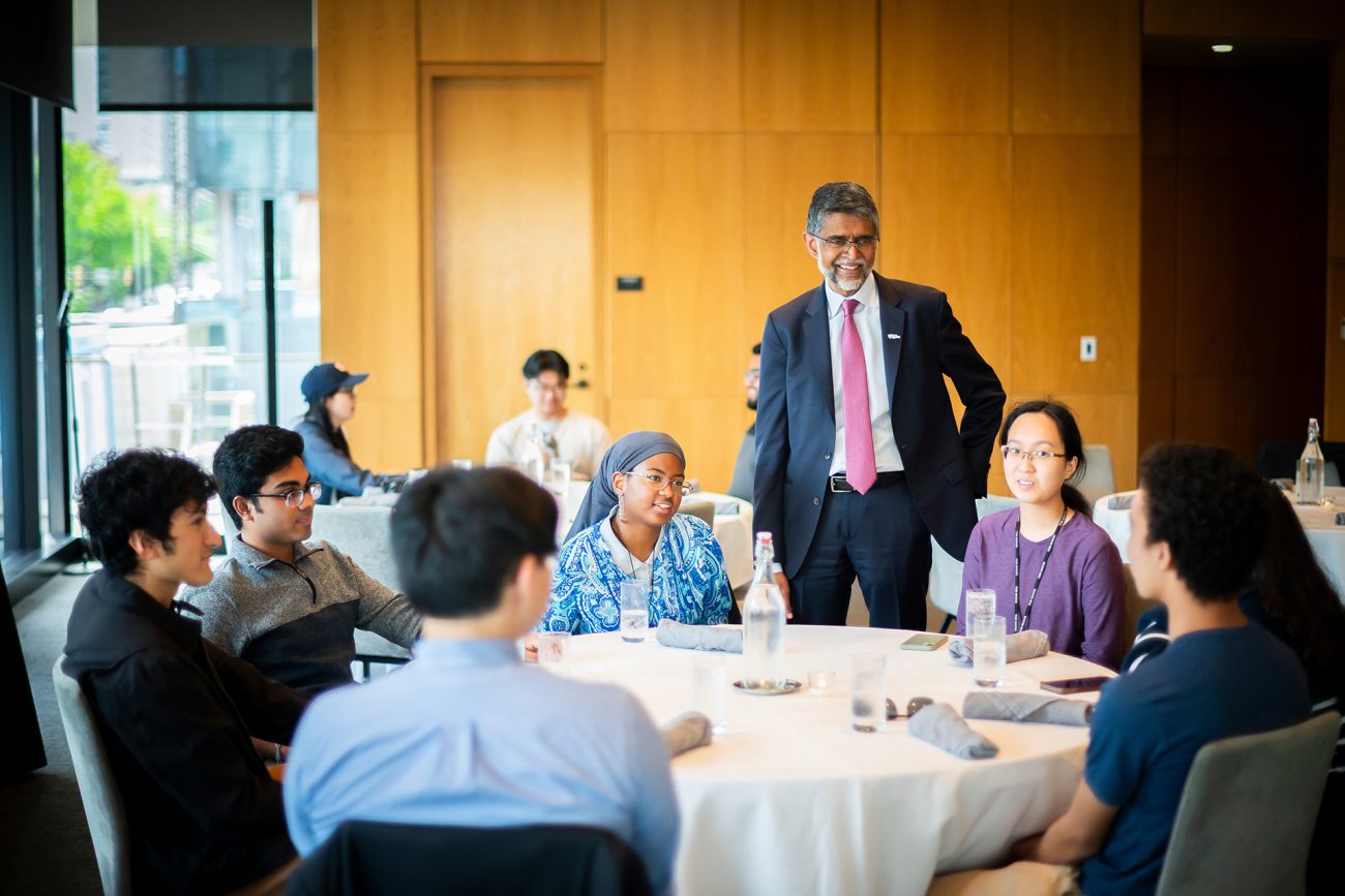 An engaging scene inside a modern meeting room with large windows, featuring a group of diverse young students sitting around a table. Penn Engineering Dean Vijay Kumar stands at the center, smiling as he interacts with the students.