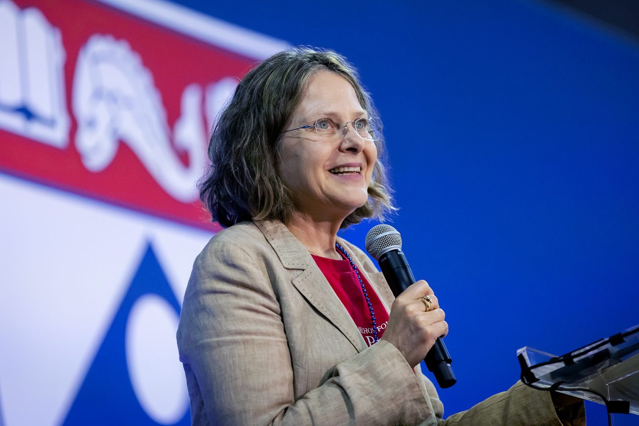 Sigal Ben-Porath, dressed in a casual blazer and a red blouse, smiles as she addresses an audience, microphone in hand.