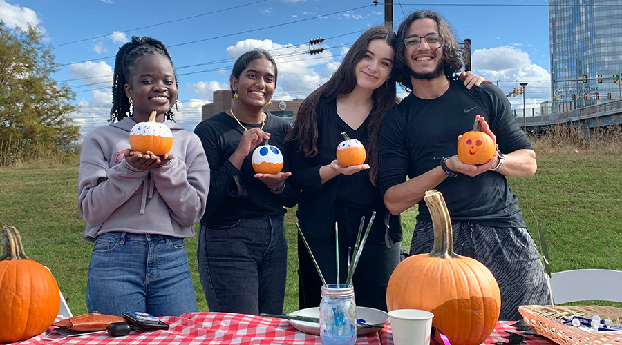A cheerful group of four diverse young adults holding painted pumpkins during a pumpkin decorating event.