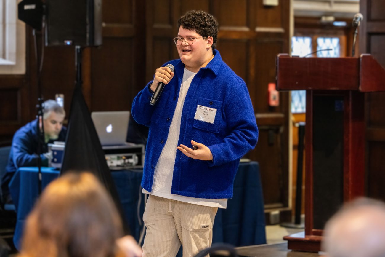 A young male speaker in a blue fleece jacket holding a microphone at a university event. He is engaging with the audience, mid-speech, smiling and gesturing with one hand.