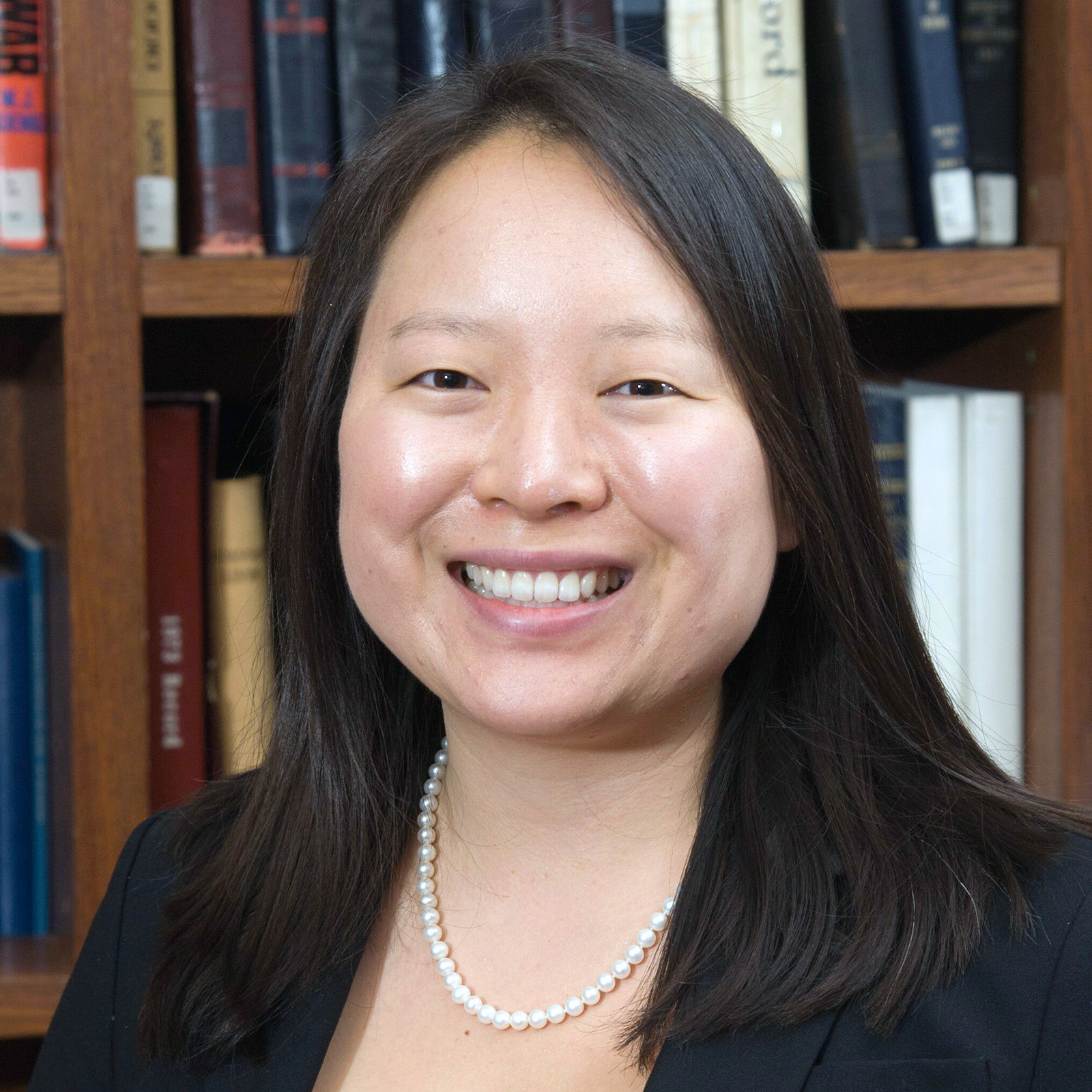 Portrait of a cheerful woman with shoulder-length black hair, wearing a black blazer and a pearl necklace, smiling in front of a bookshelf filled with various books.