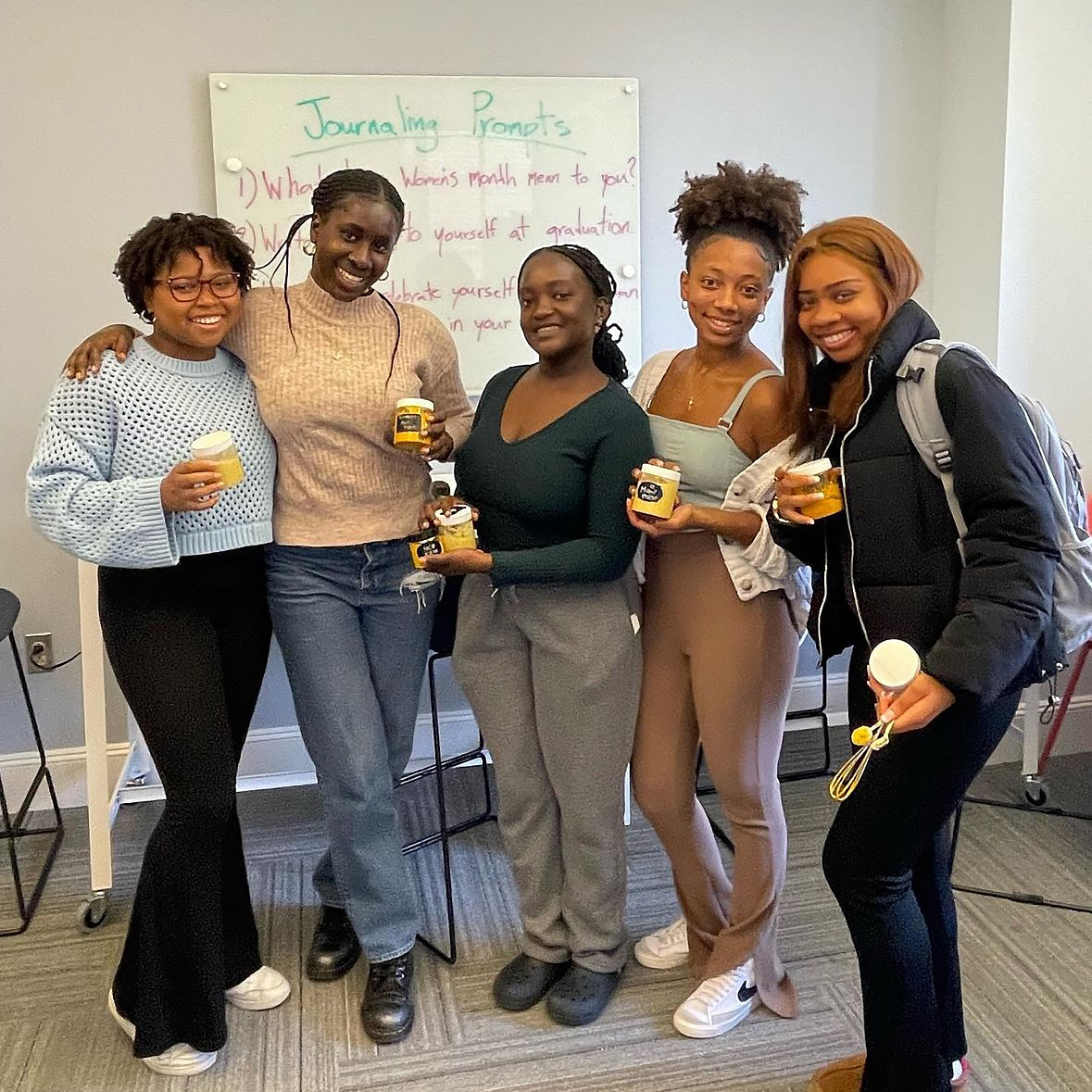 Five joyful young women of diverse backgrounds stand together holding mason jars, smiling in front of a whiteboard with journaling prompts written on it.