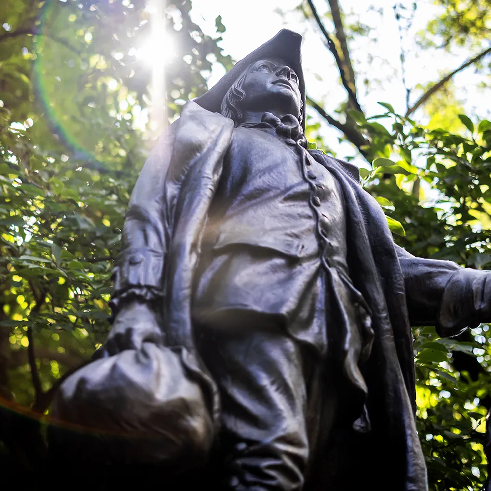 A bronze statue depicting a young Benjamin Franklin wearing a tricorn hat and colonial-era clothing, holding a satchel in his hand.